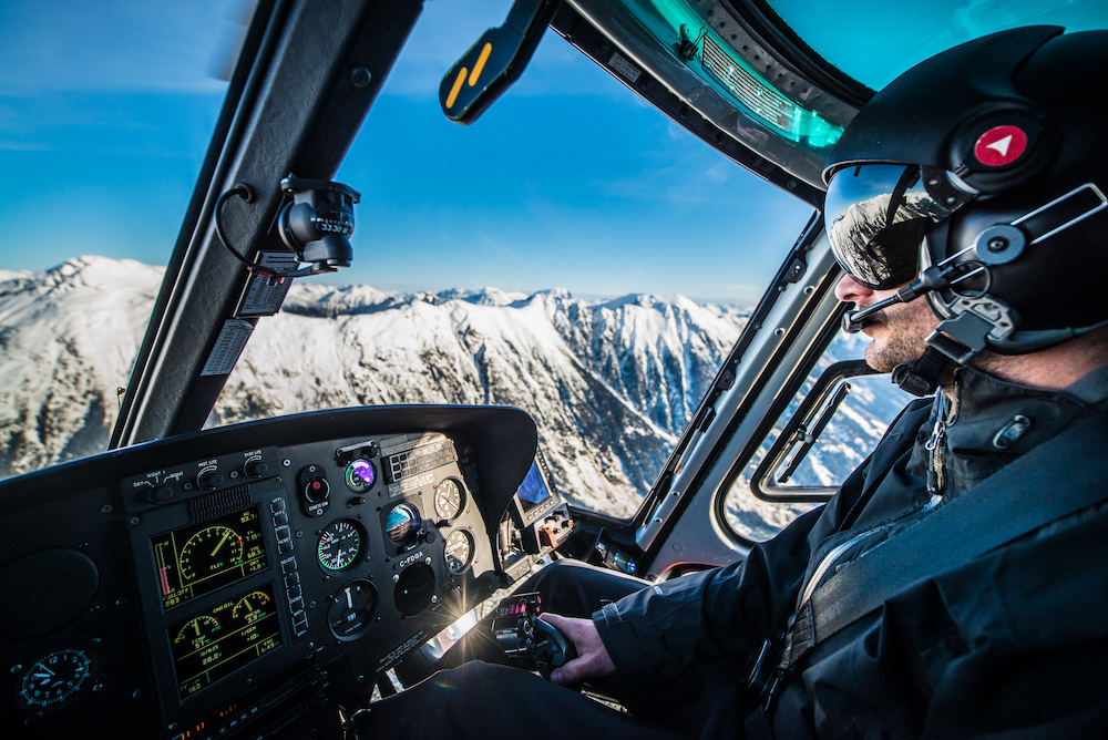 Inside view of helicopter and pilot flying over mountains in British Columbia.