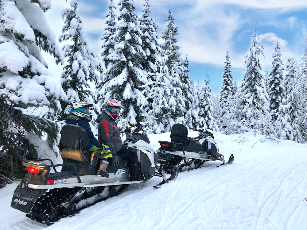 Two people on snowmobile in snowy landscape with pine trees on snowmobile safari in Whistler.